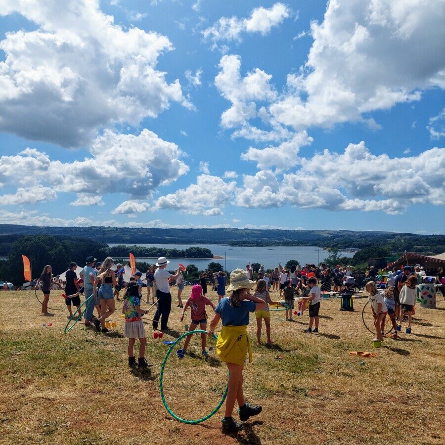 Girl with a hat holding a green hula-hoop and other children with a variety of toys, on a grassy hillside over looking a large lake and a blue sky with little fluffy clouds.