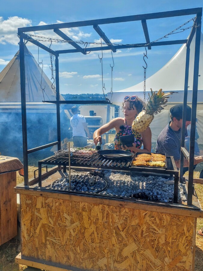 Woman cooking fish and potatoes rosti over a large charcoal barbeque. A pineapple hangs above the fire